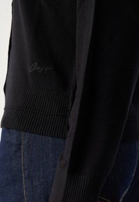 Svart strikket genser med ribbestrikkede mansjetter og kant. Har et subtilt logo på siden og er parret med mørke jeans med synlig søm.