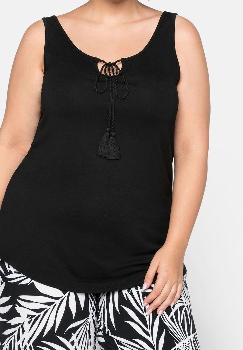 Haut noir sans manches avec un laçage devant et des pompons, porté avec un pantalon à motif feuilles tropicales noir et blanc.