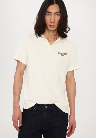 Kortärmige, crèmekleurige polo met een zachte textuur, voorzien van een blauw-rood "POLO SPORT" logo op de linkerborst. Gekenmerkt door een combinatie met donkere jeans.