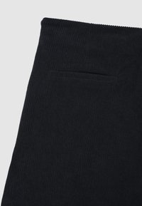 Schwarzer Cordstoff mit einer gerippten Struktur, versehen mit einem Bund und einer einzigen Gesäßtasche auf der rechten Seite.