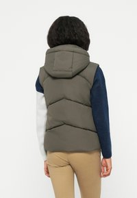 Khaki gepolsterte Weste mit Kapuze, ausgestattet mit einer Chevron-Steppung und einem hohen Kragen. Kombiniert mit einem blau-weißen Langarmpullover und beigen Hosen.