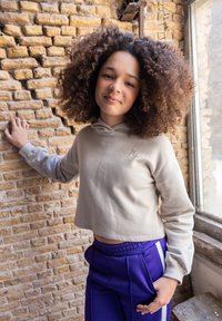 Lichtgrijze, geknipte hoodie met een capuchon en een voorzak, gecombineerd met paarse broek met witte strepen aan de zijkant. Achtergrond van een gemetselde, gestructureerde muur.