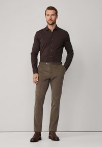 Hombre vestido con una camisa de botones marrón oscuro, pantalones oliva con ajustadores laterales, y botas de cuero marrón, de pie frente a un fondo liso.