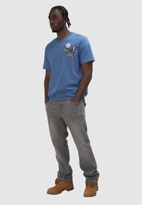 Joven con cabello trenzado, vistiendo una camiseta gráfica azul, jeans grises y botas de trabajo marrones, de pie con las manos en los bolsillos contra un fondo gris.