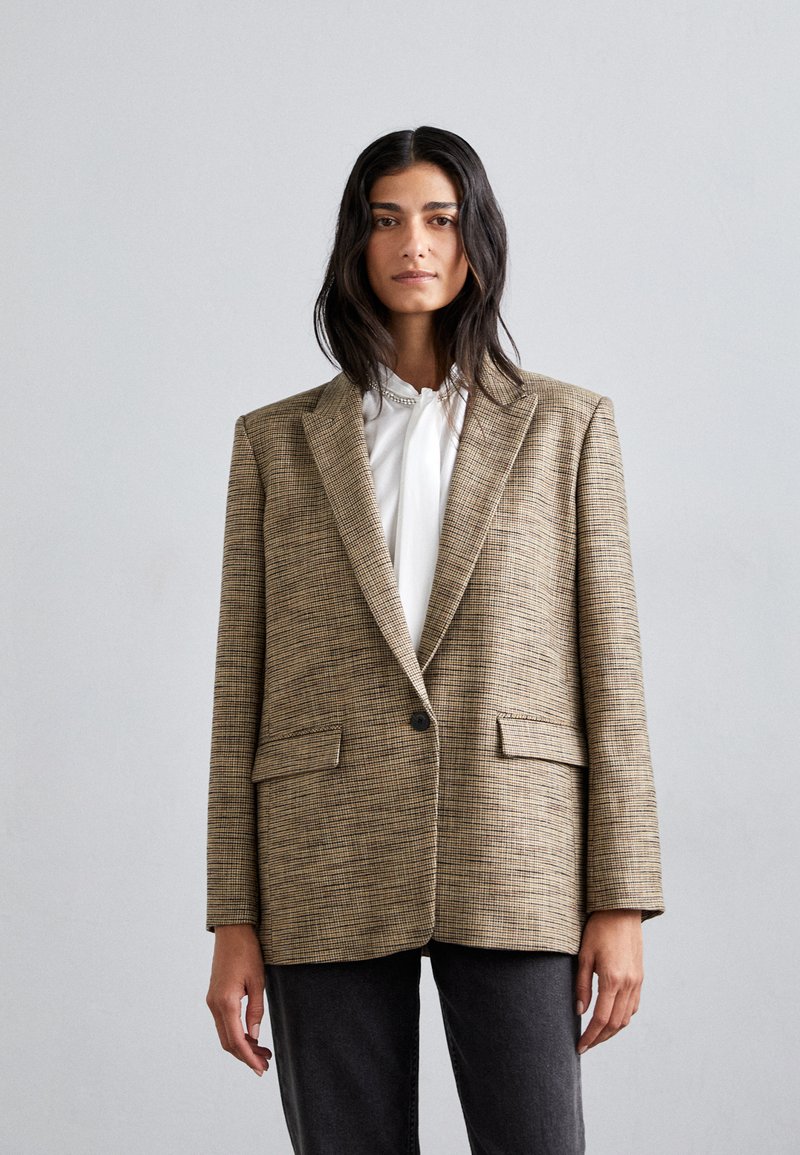 Kvinde med langt mørkt hår iført en oversized brun ternet blazer over en hvid bluse og sorte bukser, mod en ensfarvet lys grå baggrund.