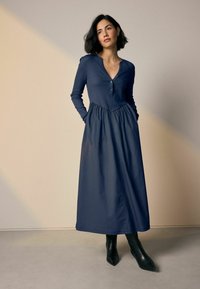 Lang marineblauwe jurk met een knoopsluiting, V-hals en lange mouwen; heeft een geraffineerde taille en zijzakken, gedragen met zwarte enkellaarzen.