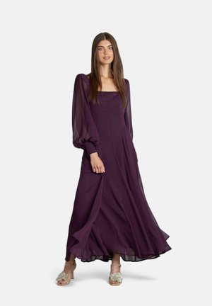 Vestido maxi morado con mangas largas y transparentes, escote cuadrado, silueta fluida y detalles plisados, combinado con sandalias de tiras embellecidas.