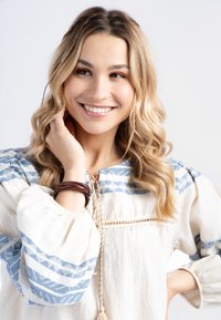 Blusa bianca con motivi geometrici blu, colletto con cordoncino, maniche a sbuffo e un bracciale intrecciato marrone. Tessuto morbido e leggero.