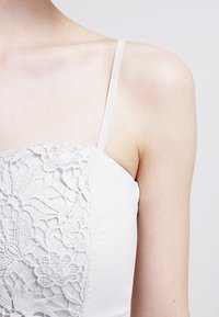 Camisole blanche avec bretelles fines ajustables, ornée de détails en dentelle délicate sur le devant, texture de tissu lisse.
