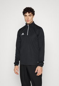 Top sportiv negru, cu fermoar la jumătate, realizat dintr-un material texturat, cu un fermoar alb și un mic logo pe partea stângă a pieptului, purtat cu pantaloni asortați.
