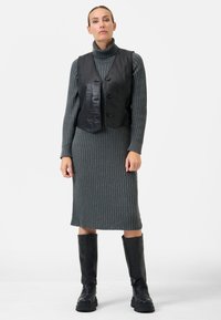 Schwarze Leder-Croptop-Weste über einem grauen gerippten Strickkleid, knielange schwarze Stiefel und ein hoher Kragen. Einfache Knopfverschluss an der Weste.