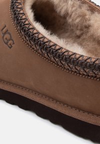 Chausson UGG marron avec extérieur en daim, doublure douce en peau de mouton, coutures décoratives au col et semelle en caoutchouc texturée.