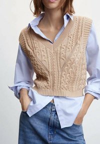 Strick ärmelloser Pullover in hellem Beige mit V-Ausschnitt und strukturierten Zopfmustern. Getragen über einem hellblauen Hemd mit Kragen und Jeans.