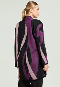 Oversized zwarte gebreide cardigan met abstracte paarse en lichtroze patronen. Zachte textuur en ontspannen pasvorm, met een kraag en lange mouwen.