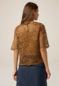 Top in pizzo di colore marrone metallico, con una texture semi-trasparente, aperture a chiave sul retro e maniche a tre quarti. Abbinato a una gonna blu.
