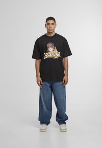 Svart bomull t-skjorte med grafisk trykk i gult, rødt og grønt. Løs passform, kortermet, kombinert med baggy blå denimjeans og joggesko.