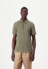 Olive rohelise polo särk, millel on kolm nupuvajutust, lühikesed varrukad ja vasakul rinnal tikandiga logo; koos heledate beežide pükstega.