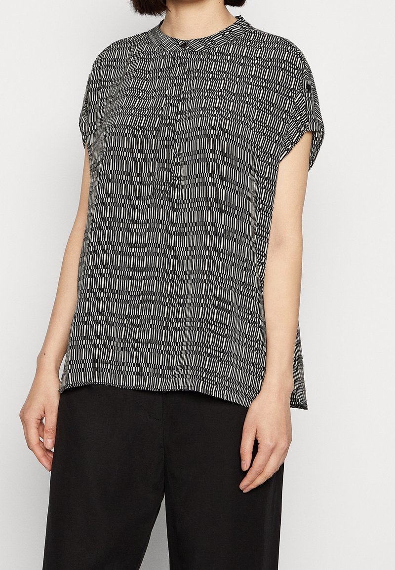 Femme portant un haut à manches courtes à motifs noir et blanc et un pantalon noir ample sur un fond uni.