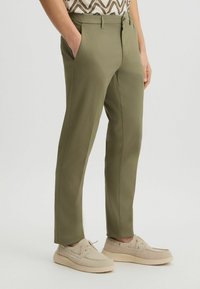 Olivgrüne Hosen mit geradem Schnitt, seitlichen Taschen und Reißverschluss. Kombiniert mit beigefarbenen Schuhen mit Schnürdetails.
