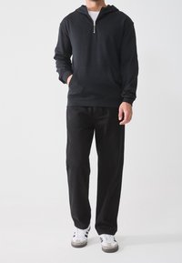Sweat à capuche noir zippé avec une poche kangourou, associé à un pantalon noir et des baskets blanches avec des accents noirs et gris.