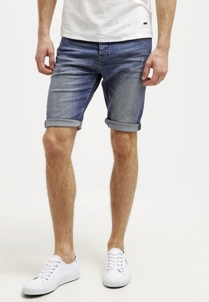 Farkkuhousut, keskipitkä sininen, käännetyt lahkeet, normaali istuvuus, yhdistettynä valkoiseen t-paitaan ja valkoisiin lenkkareihin, esittelee rentoa kesätyyliä.