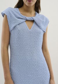 Rochie de un albastru deschis, cu un material texturat, decolteu în V și detaliu în formă de fundiță răsucită pe umeri. Finisaj neted cu un aspect moale și ușor crăpat.