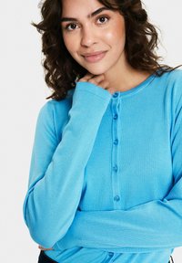 Vaaleansininen ribbikuvioinen neuletakki, jossa on pyöreä pääntie, pitkät hihat ja viisi samanväristä nappia edessä. Pehmeä tekstuuri.