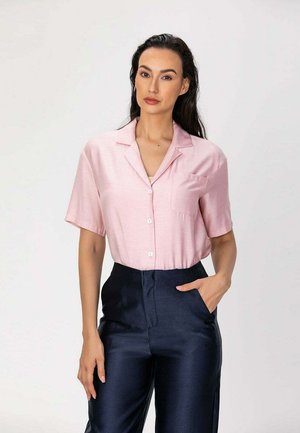 Femme aux longs cheveux foncés porte une chemise rose clair à manches courtes boutonnée et un pantalon taille haute bleu marine foncé, debout avec une main dans la poche.