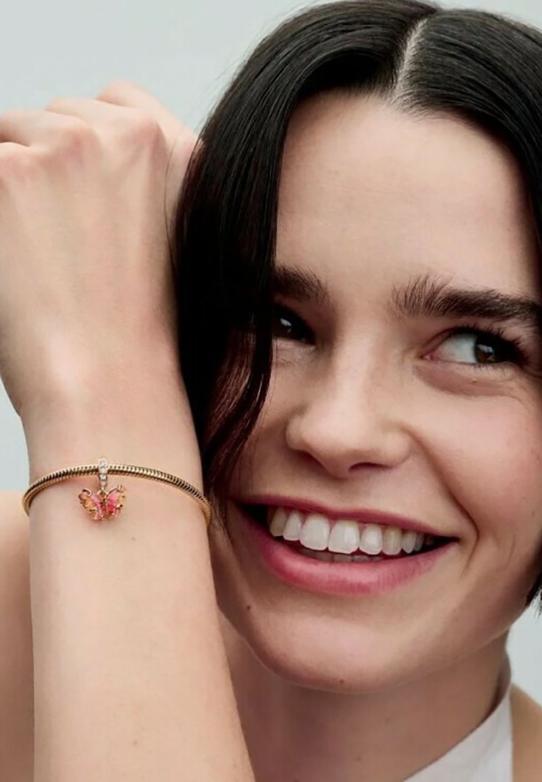 Zlati zapestnica z obeskom v obliki metulja, ki ima roza in prozorne akcentne detajle. Gladka tekstura, okrogla oblika in zapiranje s sponko.