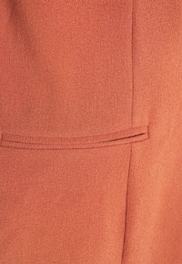 Rostfärgad tyg med en jämn textur och en subtil lyster. Har en enkel, smal ficka. Sömnadsdetaljer synliga på högra sidan.