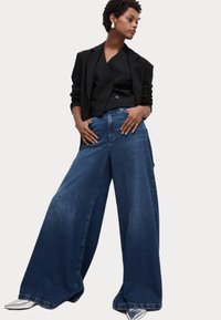 Vaqueros de denim azul de pierna ancha combinados con un blazer negro de doble botonadura y tacones plateados de punta afilada. Textura suave, diseño casual.