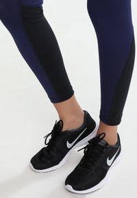 Černé sportovní boty s bílým „swooshem“ Nike, doplněné tmavě modrými a černými legínami s texturou. Detailní pohled od poloviny lýtek dolů.