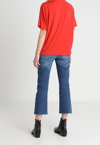 Top rosso a maniche corte abbinato a jeans cropped blu. Il modello indossa stivaletti neri alla caviglia con dettagli in frangia. Design semplice, senza motivi visibili.