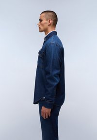 Joven de pie de lado, vistiendo una camisa de mezclilla azul oscura y jeans azules, contra un fondo ligero y plano.