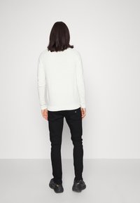 Person with shoulder-length dark hair stands facing a plain white wall, wearing a white sweater, black pants, and black sneakers.