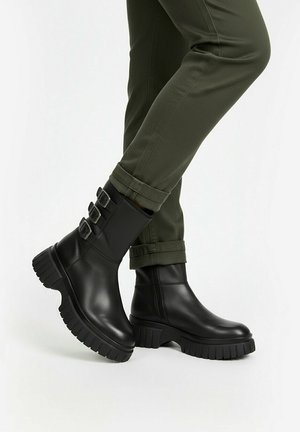 Person trägt schwarze Leder-Stiefeletten mit dicken Sohlen und drei Schnallenriemen, kombiniert mit hochgekrempelten olivgrünen Hosen.