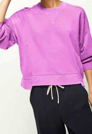 Persoon draagt een loszittend paarse trui met geribbelde manchetten en zoom, gecombineerd met een zwarte broek met een witte trekkoord.