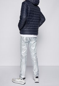 Polstret navy jakke med hætte, metalisk sølvbukser og hvid skjorte, komplet med hvide og grå sneakers. Glatte teksturer, moderne design.