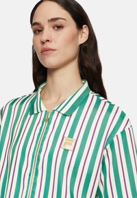 Chaqueta a rayas verdes y blancas con acentos morados, cierre frontal con cremallera y cuello. Presenta un logo dorado en el pecho. Textura suave.