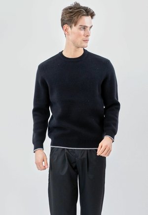 Jeune homme portant un pull noir à col rond et un pantalon noir, debout et regardant sur le côté sur un fond clair uni.