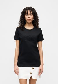 Μαύρο μπλουζάκι με κοντά μανίκια, στρογγυλή λαιμόκοψη και ανάγλυφο σχέδιο λογότυπου. Συνδυάζεται με λευκή φούστα με χρυσά διακοσμητικά κουμπιά.