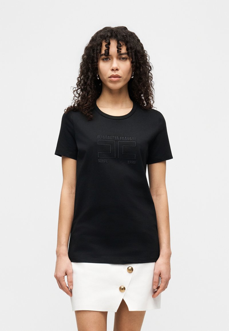 Μαύρο μπλουζάκι με κοντά μανίκια, στρογγυλή λαιμόκοψη και ανάγλυφο σχέδιο λογότυπου. Συνδυάζεται με λευκή φούστα με χρυσά διακοσμητικά κουμπιά.