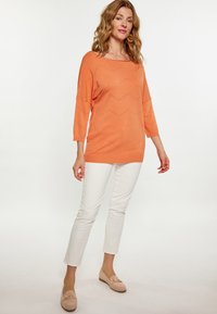 Pomarańczowy sweter dzianinowy w zygzakowy wzór, rękawy o długości trzech czwartych. W połączeniu z białymi spodniami slim-fit i jasnymi mokasynami.