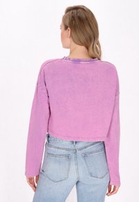 Top lungo a maniche lunghe di colore viola, tagliato in vita, con una texture morbida e collo a giro. Presenta spalle cadenti e una vestibilità rilassata. Sono visibili jeans di un lavaggio chiaro.