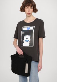 Zwarte gebreide handtas met een textuur oppervlak en dubbele handvaten, vastgehouden in een hand. De tas heeft een metalen label met tekst eraan bevestigd.