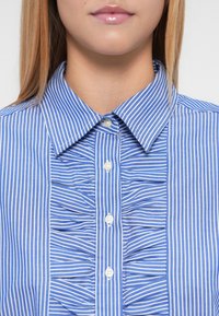 Μπλε και λευκό ριγέ πουκάμισο με γιακά με κουμπιά, μπροστινή πτυχωτή παντούλα και εμφανή λευκά κουμπιά στο κέντρο.