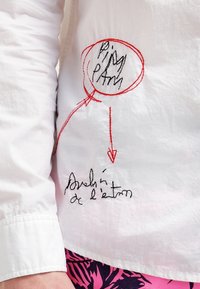 Vit skjorta med broderad text i svart och rött. Cirkulär design med orden "Ping path" och en pil som pekar nedåt.