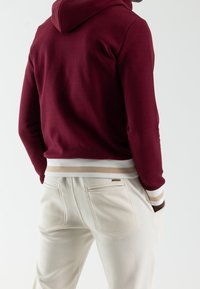 Bordowa bluza z kapturem o fakturowanym wykończeniu, z białymi i beżowymi prążkowanymi mankietami oraz ściągaczem, w komplecie z kremowymi spodniami.