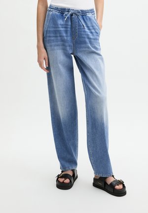 Femme portant un pantalon en jean bleu clair délavé à cordon et des sandales à plateforme noires, debout avec une main dans la poche.