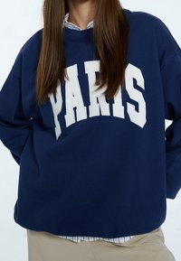 Granatowy sweatshirt wykonany z miękkiej tkaniny, z dużą białą grafiką "PARIS" na przodzie oraz luźnym krojem.